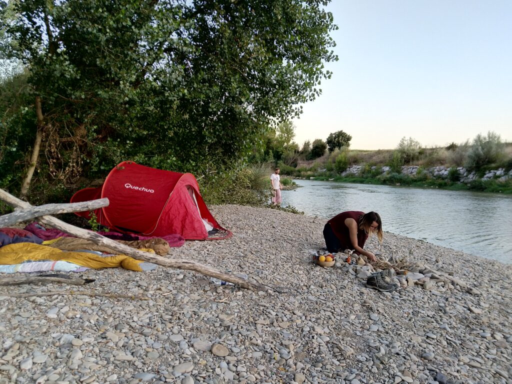 Sur un bord de rivière, une tente est installée à côté d'un campement sommaire monté grâce à des morceaux de bois. Une personne est en train de faire un feu de camp avec des galets.